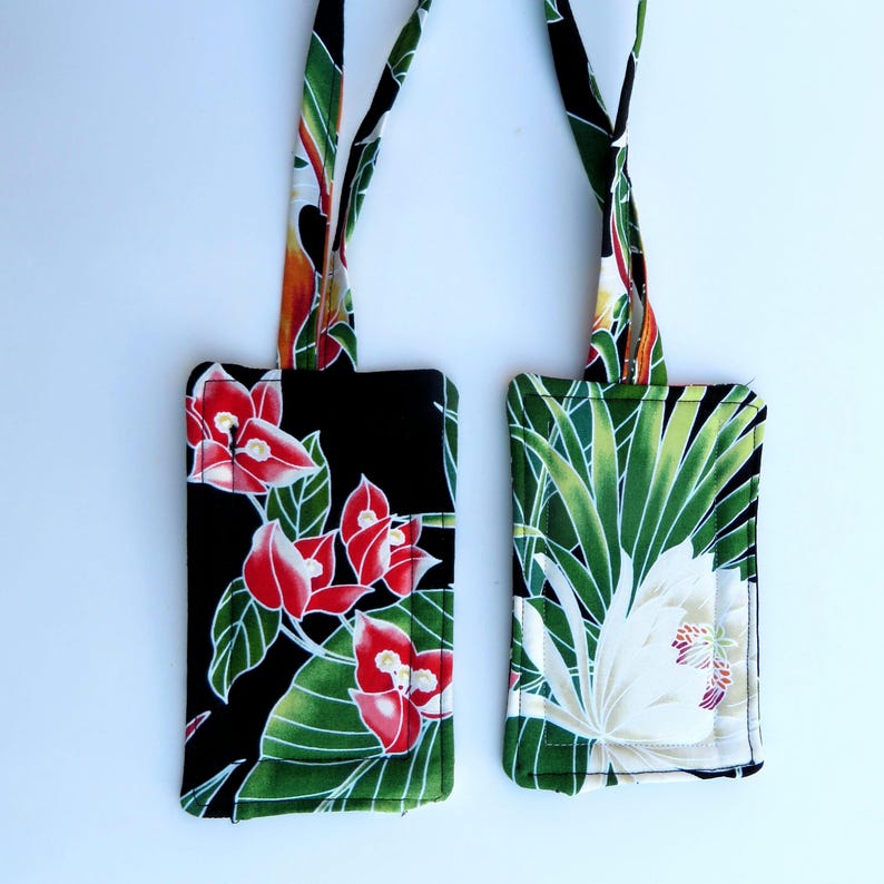 Peut inclure: Deux &eacute;tiquettes de bagages en tissu avec des motifs floraux. L'une pr&eacute;sente des fleurs rouges sur fond noir, l'autre une fleur blanche et des feuilles vertes sur fond vert. Les deux ont des sangles assorties avec un motif floral color&eacute;.