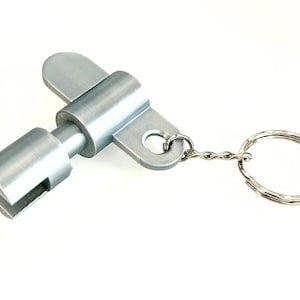 Puede incluir: Un llavero plateado en forma de llave. La llave tiene un cuerpo cilíndrico con un extremo ranurado y una parte superior plana y rectangular. El llavero está unido mediante una pequeña cadena.