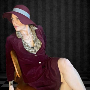 May include: A burgundy velvet suit with a matching wide-brimmed hat featuring a light blue band. The suit includes a blazer and skirt, paired with a patterned blouse and layered necklaces. The outfit is worn while seated on a wooden chair.