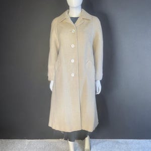 May include: A light beige, knee-length coat with a classic collar and three white buttons. The coat has long sleeves and two front pockets. The coat is displayed on a mannequin against a dark gray background.