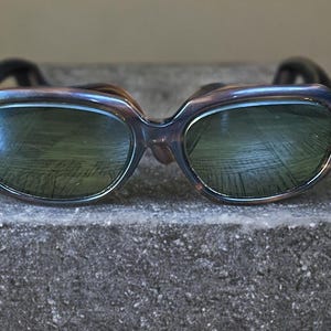 May include: Vintage sunglasses with a dark tortoiseshell frame and green-tinted lenses. The rectangular frames have a subtle cat-eye shape. The sunglasses are resting on a textured, gray surface.