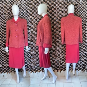 May include: A vintage coral skirt suit on mannequins. The jacket has a textured finish, a notched collar, and three buttons. The skirt is knee-length. The suit has contrasting trim on the collar and cuffs. The background has a brown and white pattern.