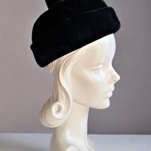 May include: A black velvet pillbox hat with a structured, rounded crown and a small, centered fabric knot detail on top. The hat features a smooth, matte texture and a clean, circular silhouette that sits firmly on the head.