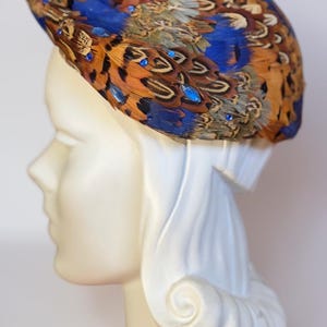 May include: A pillbox hat covered in layered brown, tan, and black patterned feathers with vibrant blue feather accents. The hat is embellished with small, round, bright blue rhinestones scattered across the surface of the feathers.