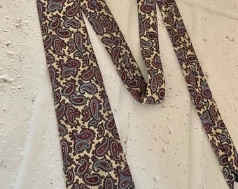 May include: A cream-colored tie with a repeating paisley pattern in burgundy, navy blue, and gray. The tie is made of a smooth fabric and is displayed against a white textured background. The paisley design adds a classic touch to this accessory.