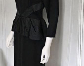 May include: A black long-sleeved dress featuring a square neckline and a fitted bodice. The waist is accented with a structured peplum detail made of a contrasting fabric, leading into a straight, knee-length skirt silhouette.
