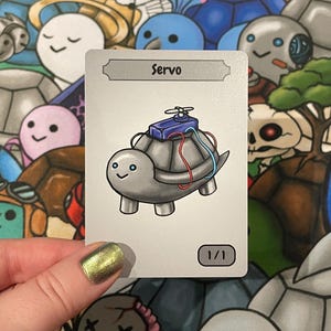 May include: A trading card depicting a cartoon turtle with a mechanical device. The card is labelled "Servo" and marked "1/1". The background is filled with various cartoon characters.
