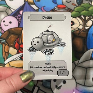May include: A trading card depicting a cartoon drone turtle with a grey shell and propellers. The card is labelled "Drone" and "Flying" with descriptive text below. The card is held against a colourful background of cartoon characters.