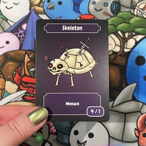 May include: A trading card featuring a cartoon skeleton turtle with a sword through its shell and arrows. The card reads "Skeleton" at the top and "Menace" at the bottom, with a number 4/1 in the corner. The background is a colorful illustration.