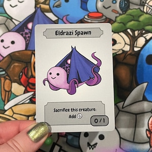 Peut inclure: Une carte à collectionner avec une illustration d'une créature Eldrazi Spawn. La carte a un fond gris clair et le texte "Eldrazi Spawn" en haut. La créature est rose avec des tentacules violets et une cape bleu foncé. Le texte "Sacrifice this creature: Add ⟳ 0/1" est également inclus.