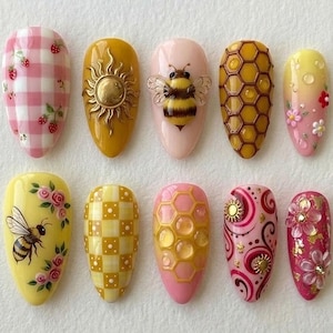 May include: A collection of ten almond-shaped artificial nails with various designs. The designs include a pink and white gingham pattern with strawberries, a gold sun, a 3D bee, a honeycomb pattern, and floral accents. Colors include yellow, pink, and gold.
