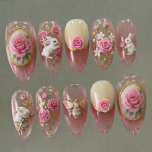 May include: A set of ten artificial nails with a pink and clear design. Each nail features 3D floral and animal designs, including roses, a bunny, and a bee. Some nails have gold accents.