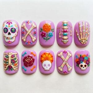May include: A set of ten press-on nails with a light purple base, featuring raised 3D designs including sugar skulls, floral arrangements, a human rib cage, a spine, a hand skeleton, and crossed bones in vibrant colors like orange, red, and teal.