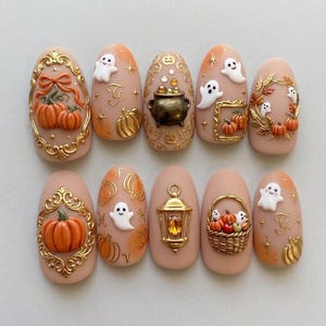 May include: A set of ten press-on nails featuring a nude base with 3D Halloween-themed embellishments. Designs include gold ornate frames, miniature pumpkins, white ghosts, a black cauldron, a gold lantern with a yellow gem, and a woven basket.