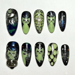 May include: A set of ten press-on nails featuring a space theme with black glitter backgrounds, light green alien cat illustrations, a silver disco ball pattern, and raised 3D accents including small spheres and a metallic UFO shape.