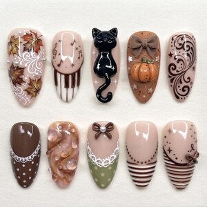 May include: A set of ten press-on nails featuring various designs including autumn leaves, piano keys, a 3D black cat, a 3D pumpkin with a bow, brown swirls, white polka dots, lace patterns, stripes, and small bows on a neutral beige base.