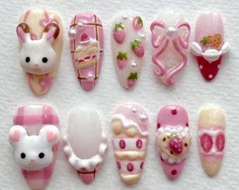 May include: A set of ten artificial nails with various designs. The nails feature a bunny, cake, strawberries, and bows in shades of pink, white, and red. Some nails have 3D elements, and others have pearl accents. The nails are oval-shaped.