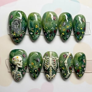 May include: Set of ten green and white press-on nails with a glossy finish. Each nail features a unique design, including a tombstone, butterflies, flowers, and a skeleton. The nails are oval-shaped and arranged on a clear display.