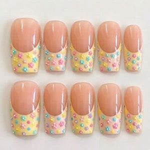 Può includere: Un set di dieci unghie finte in stile french manicure con base color carne e punta giallo pallido. Ogni unghia è decorata con piccoli dettagli floreali in rilievo multicolori nelle tonalità del rosa, blu e giallo sulle punte.