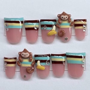 May include: A set of ten press-on nails, each with a pink base and decorated with brown, turquoise, and yellow stripes. Some nails feature a 3D monkey and banana design, plus small rhinestones. A playful nail art set.
