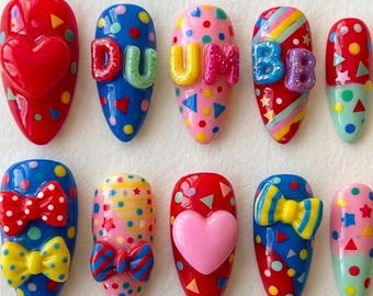 May include: A collection of ten colorful, decorated artificial nails. Designs include hearts, bows, polka dots, and the word "DUMB" in raised letters. The nails feature a variety of colors, including red, blue, pink, and yellow, with geometric shapes.