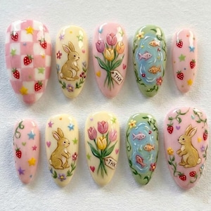 May include: A collection of ten painted artificial nails with spring-themed designs. The designs include strawberries, bunnies, tulips, fish, and floral patterns. Some nails feature the text "THX". The colour palette includes pink, yellow, green, and white.
