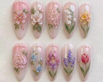 Op de afbeelding: Tien kunstnagels met een lichtroze basis, elk versierd met een ander 3D bloemmotief. De bloemen omvatten kersenbloesems, rozen en irissen, met gouden accenten en parelversieringen.