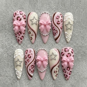 May include: A collection of ten long, almond-shaped false nails. Designs include pink leopard print, pink bows, silver star accents, and a brown and white swirl pattern. Some nails have a silver beaded border.