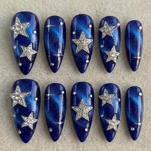 May include: Set of ten artificial nails with a dark blue galaxy design. Each nail features a silver rhinestone star and small silver accents. The nails are oval-shaped and have a glossy finish.