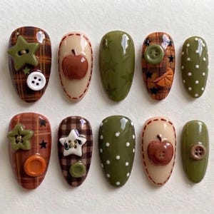 Può includere: Un set di dieci unghie finte con vari design, tra cui motivi a quadri marroni, superfici verde oliva tinta unita con pois bianchi e decorazioni 3D in rilievo come piccoli bottoni, stelle e forme di mela rossa lucida.