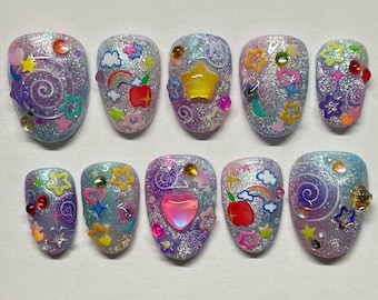 Peut inclure: Un ensemble de dix faux ongles ovales et scintillants. Chaque ongle est décoré de breloques colorées, notamment des étoiles, des arcs-en-ciel, des pommes et des cœurs. Les ongles ont une finition chatoyante et irisée avec une variété de couleurs.