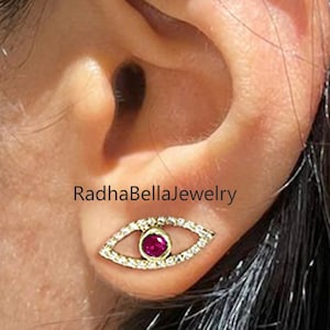 Può includere: Primo piano di un orecchio con un orecchino a lobo dorato. L'orecchino ha la forma di un occhio, con un centro rosso e piccole pietre bianche intorno. Il testo "RadhaBellaJewelry" è visibile.