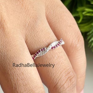 以下が含まれることがあります： 小さな赤い宝石と透明で輝く石が交互に並んだ、ひねりのあるデザインのシルバーリング。指にはめられており、「RadhaBellaJewelry」のブランド名が見えます。エレガントなデザインです。