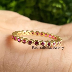May include: A gold-colored ring with a continuous row of small, round, red gemstones. The ring is held in a hand, with the text "RadhaBellaJewelry" visible on the ring. The background is a blurred green.