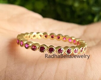 Könnte beinhalten: Ein goldfarbener Ring mit einer durchgehenden Reihe kleiner, runder, roter Edelsteine. Der Ring wird in einer Hand gehalten, mit dem Text "RadhaBellaJewelry" sichtbar. Der Hintergrund ist ein verschwommenes Grün.