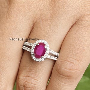 May include: A hand wearing a silver ring with a large oval ruby center stone, surrounded by a halo of smaller white gemstones. The ring is set on a silver band, and the hand is near green leaves. The text "RadhaBella jewelry" is visible.