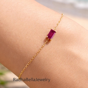 Puede incluir: Una delicada pulsera dorada con una gema rectangular de color rojo intenso. La pulsera tiene un diseño de cadena fino y elegante. El fondo muestra una escena de playa con agua azul y arena. El texto "RadhaBellaJewelry" es visible.