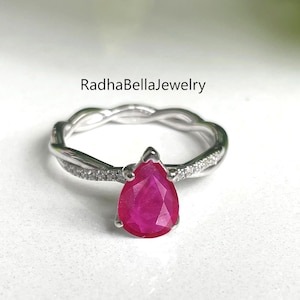 May include: A silver ring featuring a vivid red, pear-cut gemstone. The band has a braided pattern and small, clear accent stones. The ring is set against a white backdrop, with the text "RadhaBellaJewelry" visible.