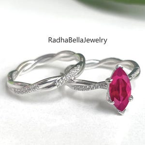 Op de afbeelding: Twee zilveren ringen. Eén ring heeft een marquise-geslepen, helderrode edelsteen. De andere ring heeft een gedraaid bandontwerp met kleine, heldere stenen. De ringen staan tegen een witte achtergrond. De tekst "RadhaBellaJewelry" is zichtbaar.