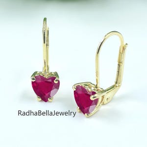 May include: A pair of gold leverback earrings, each with a heart-shaped, deep red gemstone. The earrings are set against a white background with a blurred green backdrop. The text "RadhaBellaJewelry" is visible.