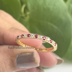 Peut inclure: Une bague en or ornée de pierres précieuses rouges et claires alternées. La bague est tenue dans une main devant un fond de feuillage vert. Le texte "RadhaBellaJewelry" est visible sur la bague.