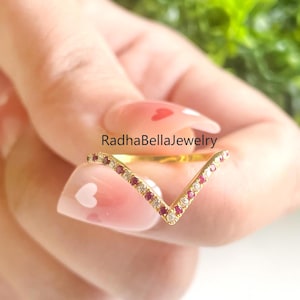 Könnte beinhalten: Ein goldfarbener, V-förmiger Ring mit einer Reihe kleiner roter und klarer Edelsteine. Der Ring wird in einer Hand mit hellrosa Nagellack und weißen Herzmustern gehalten. Der Text "RadhaBellaJewelry" ist sichtbar.