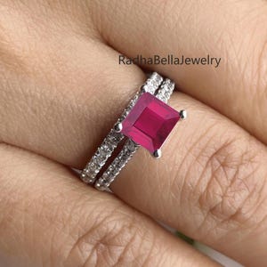 May include: A two-piece ring set featuring a square-cut, vibrant red gemstone. The band is adorned with small, clear gemstones. The rings are set in a silver-colored metal. The text "RadhaBellaJewelry" is visible.