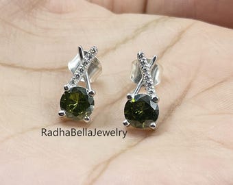 May include: A pair of silver stud earrings with olive-green oval gemstones. Each earring features a small, sparkling accent above the main stone. The earrings are displayed on a hand, with the text "RadhaBellaJewelry" visible.