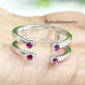 Puede incluir: Un anillo plateado con un diseño de doble banda, con cuatro pequeñas piedras rojas redondas. El anillo se sostiene en una mano, con el texto "RadhaBellaJewelry" visible.