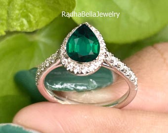 May include: A silver ring with a pear-shaped, emerald-green gemstone and a halo of small, clear stones. The band also has small, clear stones. The text "RadhaBellaJewelry" is visible. The ring is a piece of jewellery.