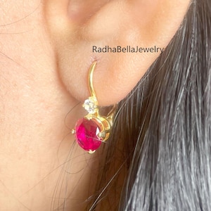以下が含まれることがあります： 丸い鮮やかな赤い宝石と小さな透明な石が付いた、金色のイヤリング。イヤリングはフープスタイルで、赤い石が耳たぶの下にぶら下がっています。「RadhaBellaJewelry」というブランド名が見えます。