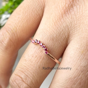 Peut inclure: Une bague en or rose avec un design incurvé, sertie de petites pierres précieuses rouges vives. La bague est présentée sur une main, avec une plante verte en arrière-plan. Le texte "RadhaBellaJewelry" est visible.
