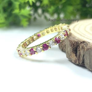 Peut inclure: Une bague d'éternité en or avec des pierres précieuses rouges et blanches alternées. La bague est posée sur une surface en bois avec un fond vert feuillu. C'est un bijou délicat.