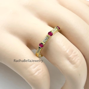 Peut inclure: Une bague en or ornée de pierres rouges et de pierres claires alternées. La bague est posée sur une main, avec le texte "RadhaBellaJewelry" visible. Un bijou délicat.